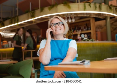 Elegant Modern Older Mature Senior Woman With Glasses Is Sitting In A Cafe With A Mobile Phone With A Cup Of Tea. The Retirement Age Is 60 Years.