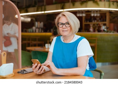 Elegant Modern Older Mature Senior Woman With Glasses Is Sitting In A Cafe With A Mobile Phone With A Cup Of Tea. The Retirement Age Is 60 Years.