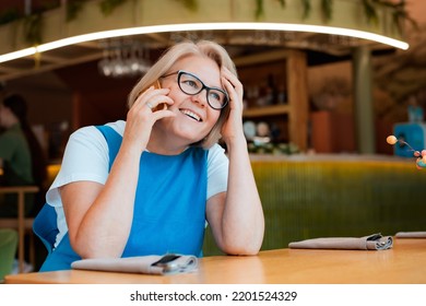 Elegant Modern Older Mature Senior Woman With Glasses Is Sitting In A Cafe With A Mobile Phone With A Cup Of Tea. The Retirement Age Is 60 Years.