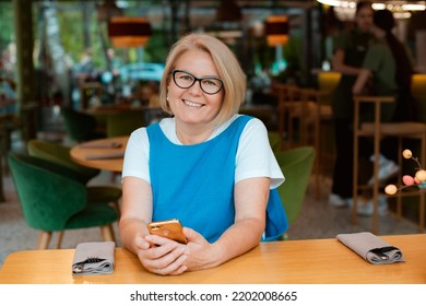 Elegant Modern Older Mature Senior Woman With Glasses Is Sitting In A Cafe With A Mobile Phone With A Cup Of Tea. The Retirement Age Is 60 Years.