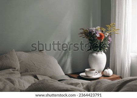 Elegant moody bedroom interior. Cup of coffee, pumpkin on retro wooden bedside table. White ceramic vase. Bouquet of dahlia, cosmos, solidago flowers. Beige muslin cushions, book in bed near window. 