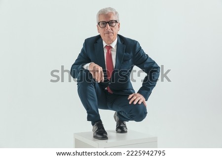 elegant old businessman with glasses holding arms on knees and crouching in front of grey background in studio
