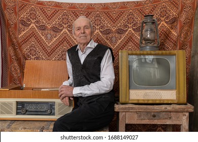 Elegant Old Man In White Shirt Sitting Between Retro Tv And Old Radio And Looking At The Camera, Ornament Wall On The Background