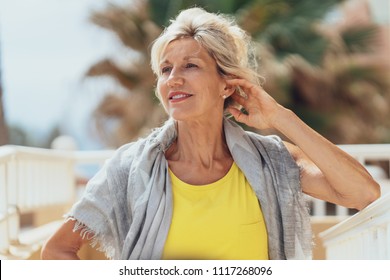 Elegant Older Blond Woman On An Outdoor Patio Posing With Her Hand To Her Hair And A Friendly Smile Looking To The Side