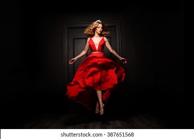 Elegant and passionate woman with tiara on her head in the red evening fluttering dress is capture in move, hovering above the ground, the wooden door is on the background
