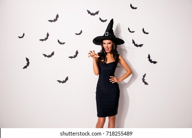 Elegant, playful, mystical female beauty. Gorgeous satanic bad fairy enchantress, hot figure, slim body, fashionable dark dress, shows teeth and arm palm with claws, stands on white background