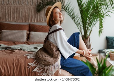 Elegant pretty woman in straw hat and white blouse posing at home, sitting on bed, morning mood. Home plants and palms. Macrame on wall. Boho style. - Powered by Shutterstock