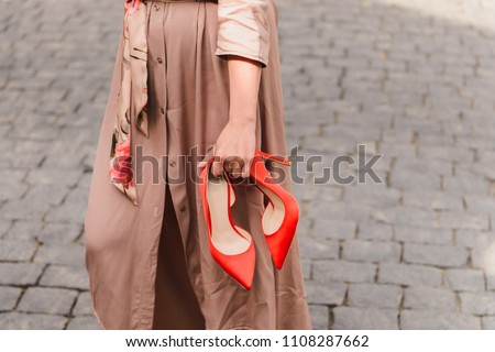 Elegant red high heels shoes in woman hands. Girl wearing beige trench coat standing on a stony pavement, holding her trendy, fashion footwear in hands