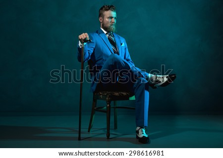 Elegant retro style gentleman with a cane sitting in a chair looking thoughtfully off to the side in a blue toned dark environment with copyspace