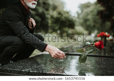 Elegant sad elderly man standing on the rain with umbrella and grieves at the grave of a loved person