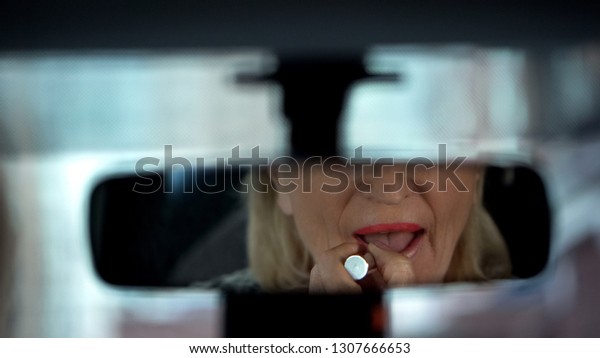 Elegant senior lady preparing for date, applying\
lipstick, looking in car\
mirror
