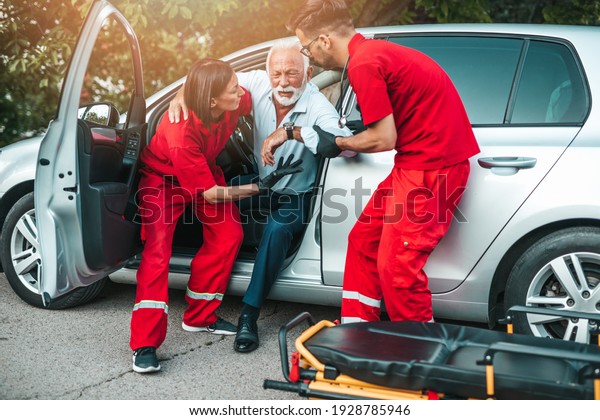 Elegant senior man with heart attack symptoms\
sitting in the car, emergency medical service workers trying to\
help him. Driver assistance service.\
