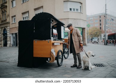 Elegant Senior Man Walking His Dog And Buiyng Street Food Outdoors In City.