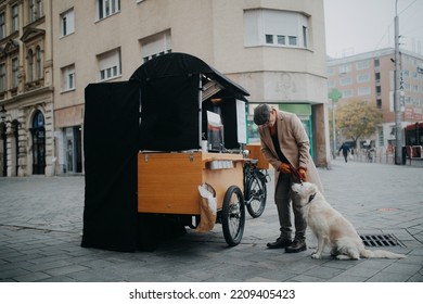 Elegant Senior Man Walking His Dog And Buiyng Street Food Outdoors In City.