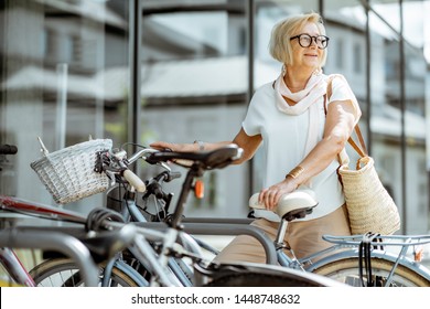 Elegant Senior Woman Parking A Bicycle Near The Modern Building Outdoors. Concept Of An Active Lifestyle On Retirement Age