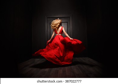 Elegant and sexy woman in the red evening fluttering dress is capture in move, running away from the ceremony (the 	
ball), the wooden door is on the background