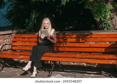 An elegant slender aged woman with long blond hair, in a black long dress, remotely works on a tablet, sits on a bench in a city park, on a bright sunny day. - Powered by Shutterstock