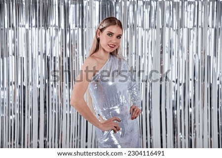 Elegant smiling young woman in glitter dress at bright tinsel background celebrating festive holiday, looking at camera. Beautiful positive charming lady model posing and present. Copy text space