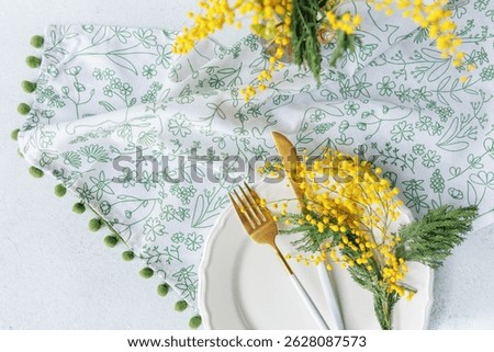 Elegant spring table decor with a white plate, gold cutlery, and fresh mimosa flowers on a floral napkin, perfect for festive occasions.