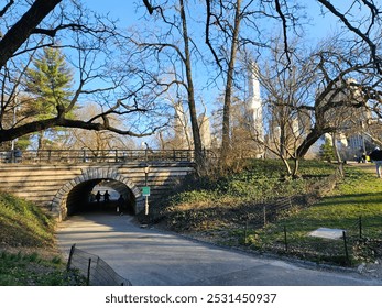 An elegant stone arched bridge forms a tunnel for a walking path in a bustling city park, where bare tree branches frame the urban skyline and warm sunlight casts long shadows on a serene early spring - Powered by Shutterstock