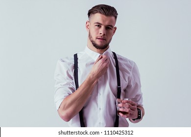 Elegant and stylish man in classical wear holding glass with wiskey in one hand and adjusting collar with another while standing against gray background. Fashionable man in white shirt and suspenders - Powered by Shutterstock