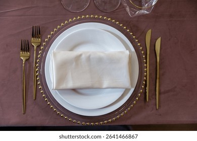Elegant table setup featuring a white plate with a neatly folded napkin placed on a decorative charger. Gold cutlery is arranged on both sides, creating a sophisticated dining presentation.
