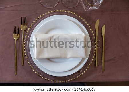 Elegant table setup featuring a white plate with a neatly folded napkin placed on a decorative charger. Gold cutlery is arranged on both sides, creating a sophisticated dining presentation.
