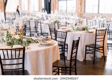 Elegant tablecloth tables and wooden chairs decorated for a wedding reception. The tables have plates, greenery, napkins, and candlesticks.