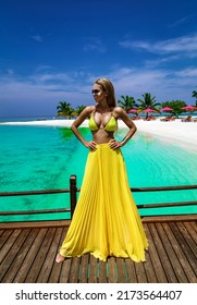 Elegant Tanned Woman In Yellow Bikini And Long Skirt On Tropical Beach On Maldives Island. Summer Fashion Model Is Walking On The Beach. Maldives  Landscape. Luxury Travel.