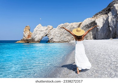 A elegant tourist woman in a white summer dress enjoys the beautiful beach of Lalaria, Skiathos island, Greece, during sunset time