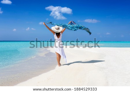 A elegant tourist woman in white summer dress walks on a tropical beach with a waving scarf in her hand