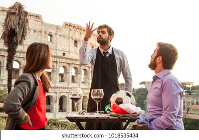 Elegant Waiter Serving Football With Italian Flag To Man Of Romantic Young Couple Sitting At Restaurant Table In Front Of Colosseum In Rome At Sunset. Girl Is Annoyed