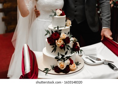  Elegant wedding cake decorated with burgundy roses and green with bride and groom on background. Wedding ceremony. Beautiful pie on the table. Brides cut cake together. Close up - Shutterstock ID 1633148320