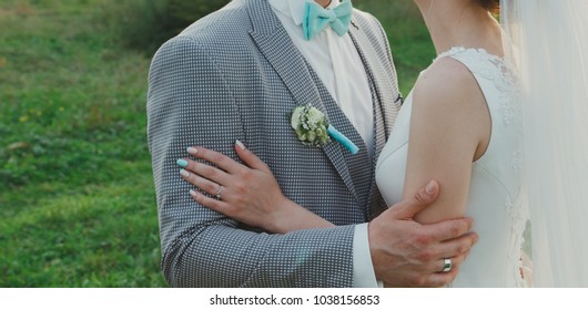 Elegant Wedding Couple Is Hugging Outdoors. Groom In Checkered Grey Suit And Tiffany Light Blue Bow Tie. Bride In Satin White Dress. Summer Romance. Wedding Buttonhole And White Golden Rings.