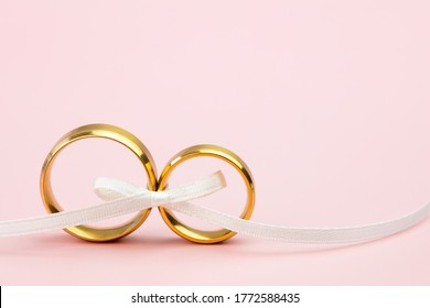 Antecedentes elegantes de boda o compromiso - un par de anillos de boda dorados y una suave cinta blanca de arco sobre el fondo rosa suave, copia espacio para el texto