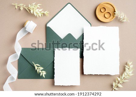 Elegant wedding stationery set. Wedding invitation cards templates, green envelopes, silk ribbon, golden rings on pastel beige background. Flat lay, top view, copy space.