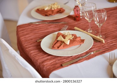Elegant wedding table setting featuring a terracotta runner, white plate with a decoratively folded napkin tied with ribbon and dried flowers, wine glasses, and silverware, with another place setting visible in the background.