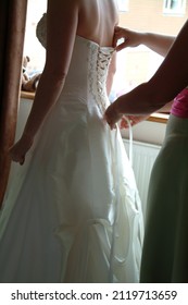 Elegant White Bridal Gown Being Laced Up From Behind. Beautiful Wedding Dress Of Silk And Lace With Bride Being Assisted With Fitting.