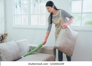 Elegant Wife In Rubber Protective Gloves Using Feather Duster Cleaning The Couch. Young Housewife Dusting Sofa Holding Up The Pillow In Bright Living Room Standing Next To The Window.