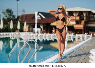Elegant woman in the black bikini on the sun-tanned slim and shapely body is posing near the swimming pool, wearing hat and stylish sunglasses