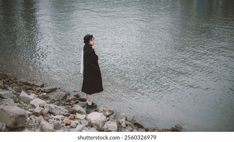 Elegant Woman in Black Coat and Hat Gazing at Serene Waterfront on a Rocky Shoreline - Peaceful Outdoor Scene. - Powered by Shutterstock