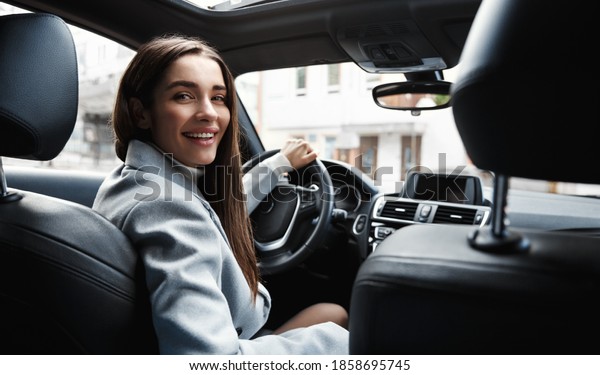 Elegant woman driver looking at\
backseat, smiling happy, asking passanger fasten seatbelt.\
Businesswoman talking to person in her car, driving at\
work.
