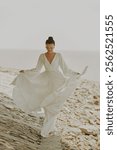 Elegant Woman in Flowing White Dress Walking Gracefully by the Seashore, Capturing Serene Beauty and Tranquility on a Sunny Day.