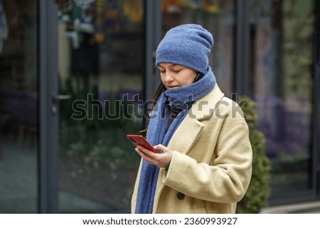 Elegant woman in knitted hat and scarf in beige coat sending text message using smartphone outside in city in winter.