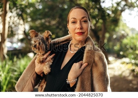 Elegant woman looking at the camera while holding her puppy outdoors. High class woman standing alone in a park during the day. Female pet owner going for a walk with her dog.