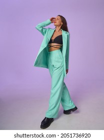 Elegant woman in mint costume blazer and pants and black top with stripes and boots standing and posing at purple background, Full length fashion portrait.