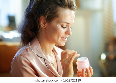 elegant woman in pajamas with pink cosmetic jar applying eye cream at modern home in sunny winter day.