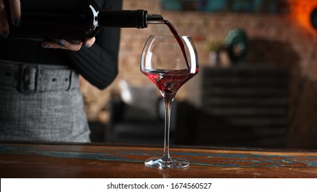 Elegant Woman Pouring Red Wine From Wine Bottle To Wine Glass At Home In A Cosy Dark Room.