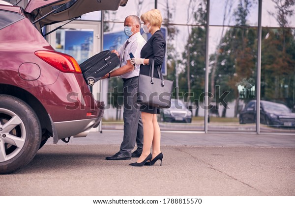 Elegant woman in protective face mask holding\
ticket and passport while gentleman getting travel suitcase out of\
car trunk