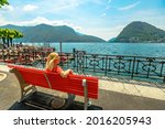 Elegant woman in a red dress enjoying Lugano city on Lugano Lake in Switzerland. Red bench by the lakefront of Lugano Lake with Monte San Salvatore mount in Ticino canton.